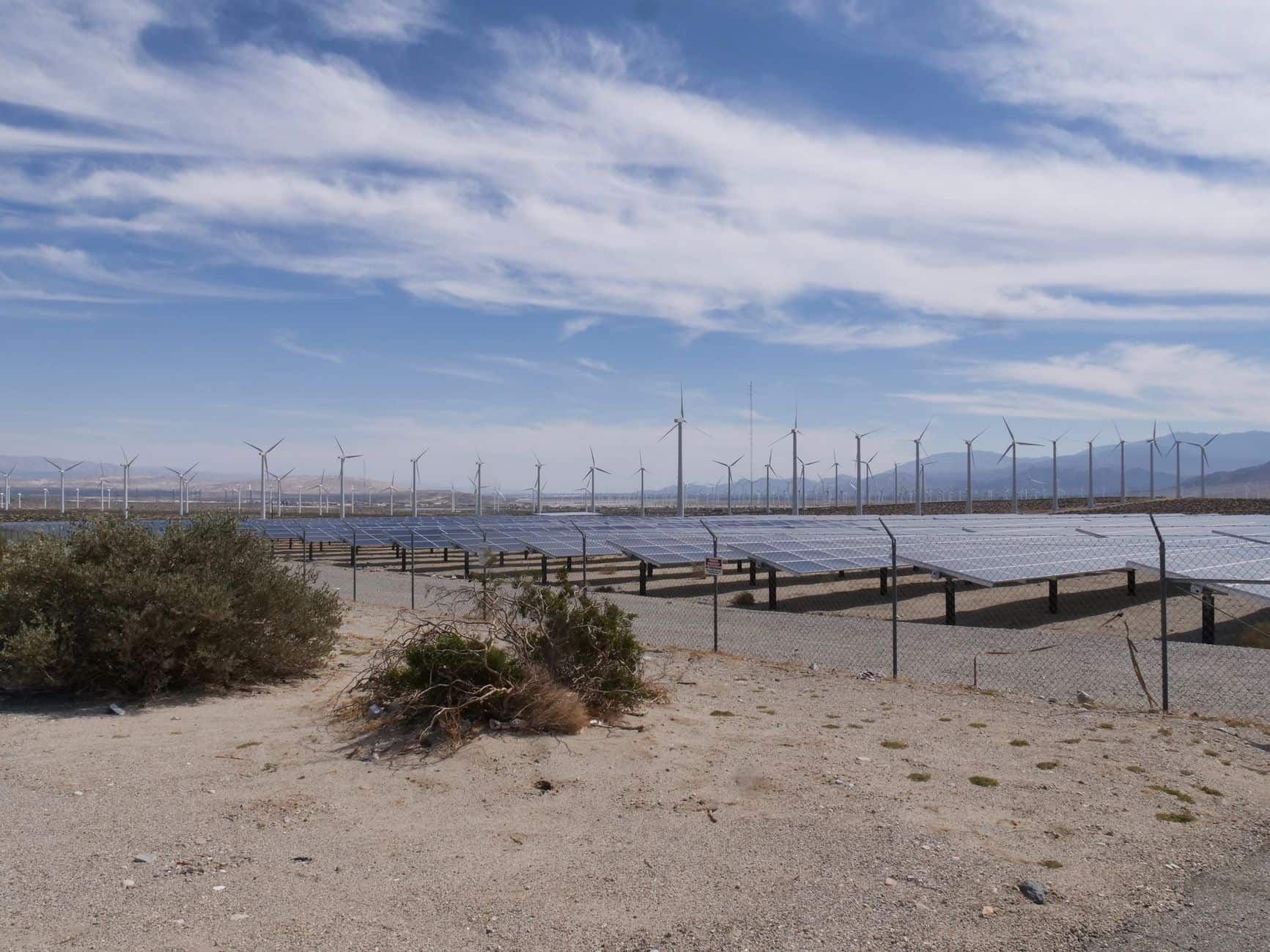 Aerial view of a vast desert solar farm with rows of panels and wind turbines under a clear blue sky