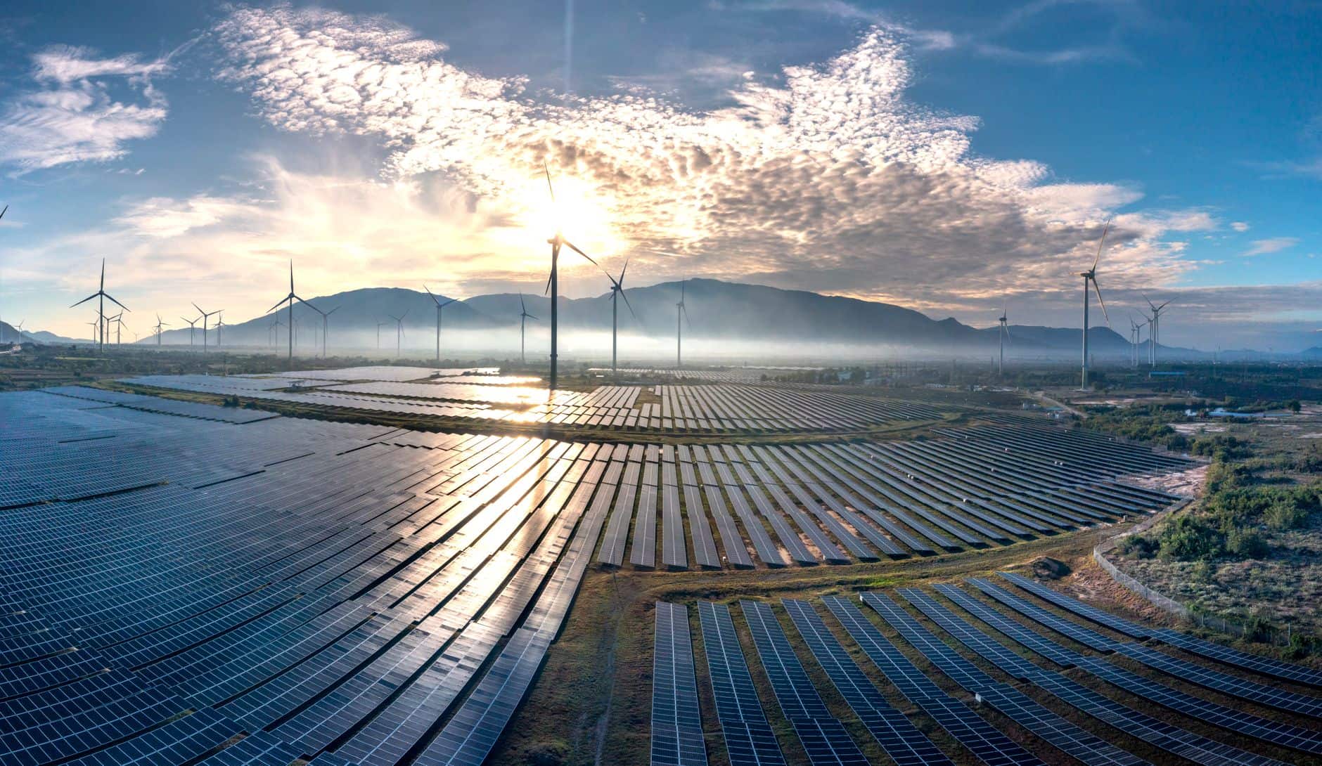Aerial view of a large solar farm alongside wind turbines at sunrise