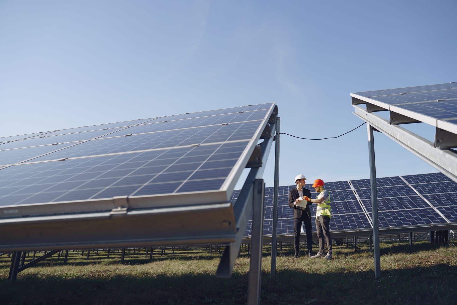 Supervisor in hard hat and formal attire discussing project paperwork beside modern solar panels under a blue sky